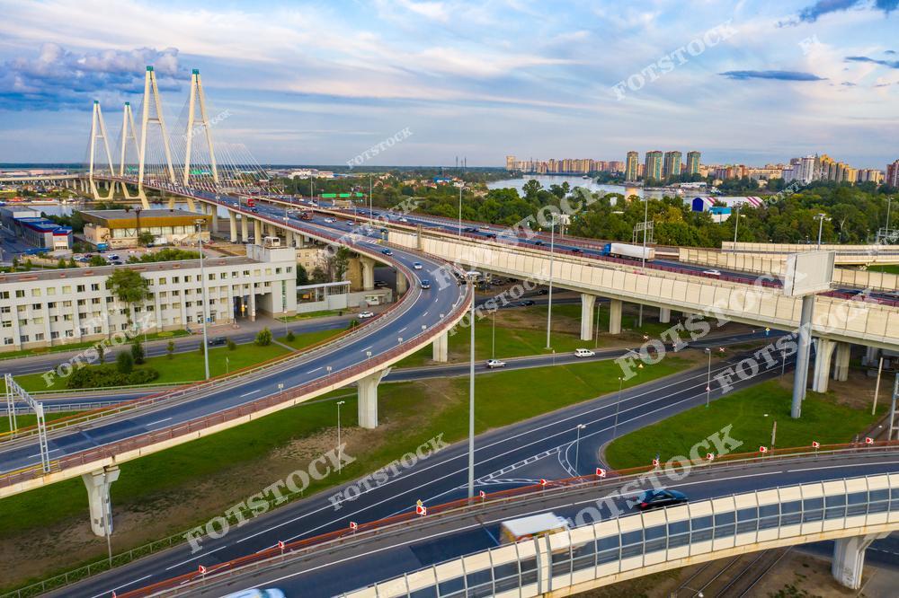 На фото изображен Вантовый мост в Санкт-Петербурге, являющийся частью Западного скоростного диаметра (ЗСД). Этот мост соединяет разные части города, обеспечивая быструю связь через Неву. На переднем плане видна сложная дорожная развязка, а вдали – зеленые зоны и многоэтажная жилая застройка. Фото передает масштаб современной городской инфраструктуры.